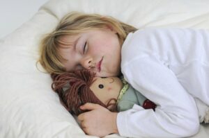 Kleines Mädchen liegt mit Puppe schlafend im Bett.